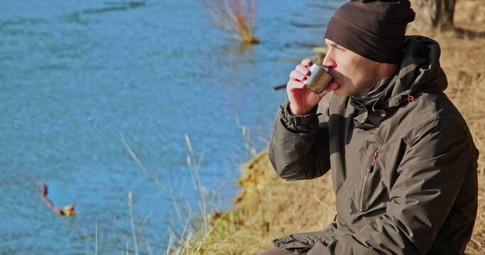 man in winter clothes on the river bank in a pine forest drinks tea and rests 