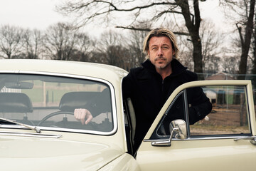 Man with blond hair and stubble in a black coat stands by a classic car in the countryside.