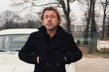 Man with blond hair and stubble in a black coat stands by a classic car in the countryside.