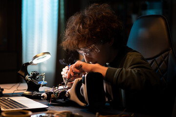 A teenager learns electronics and soldering wires on an old robot. A child works on a toy in room. There is smoke rising from the solder iron, boy is wearing safety goggles.