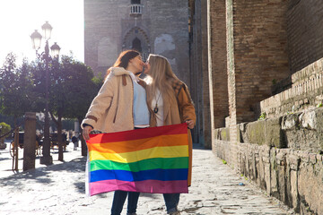 Fototapeta na wymiar Marriage of lesbians on holiday and tourism in seville. They are in front of the cathedral and they are holding the gay pride flag in their hands while kissing. Concept of equality and lgtb rights.