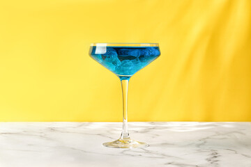 Iced blue lagoon alcohol cocktail drink on yellow background. Blue Summer beverage. Marble table.