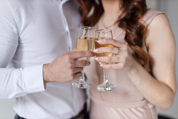 A young man in a white shirt and a girl with a ring on his hand in a beige dress drink champagne in crystal glasses, close-up without faces