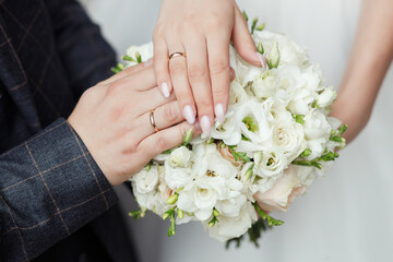 Obraz na płótnie Canvas Bride and groom in black suit and wedding dress hold hands with gold wedding rings on a wedding bouquet of white and beige roses, close-up without faces