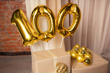 Inflatable balloons of gold color in the form of numbers represent the number one hundred, on gift boxes against a background of beige fabric