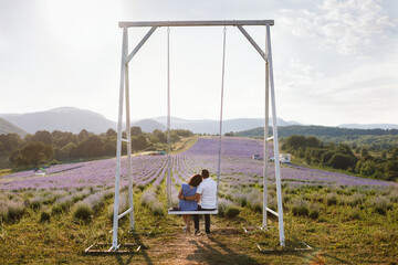 Lovers man and woman ride on a big white swing on a lavender field and watch the sunset in the mountains, film noise effect