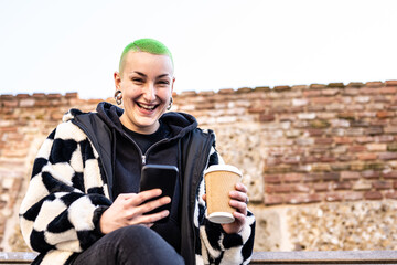 young woman with short and green colored hair smiling looking at the camera, sustainability concept of use disposable coffee cup, curvy female alternative beauty, copy space on brick wall background - Powered by Adobe