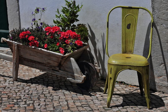 Street decoration-red azalea and violet flowers-wooden wheelbarrow-lemon yellow chair. Tavira-Portugal-088