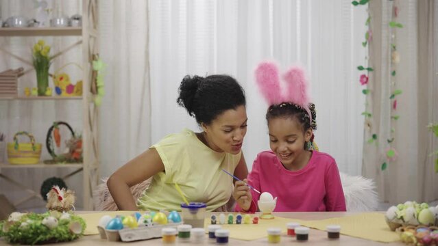 Cute little girl with funny bunny ears decorates eggs with brush and paints. African American mom and daughter are sitting in festively decorated room at home. Happy easter. Slow motion ready 59.97fps