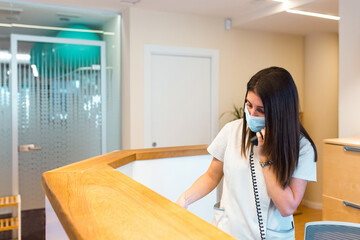 Female receptionist with a face mask attending at a call while working at the front desk of a health center.