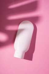 Plastic white tube for cream or lotion. Skin care or sunscreen cosmetic in top view on pink background with palm leaves shadow. Beauty concept for face care
