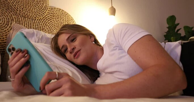 Young woman lying down on her bed floor using a smartphone at night inside her bedroom apartment. High quality video. Mobile phone, technology, concept. 