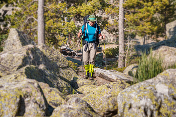 Male traveler with trekking poles and a large backpack walks along a trail among huge stones and boulders in a national park