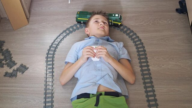 a teenager is playing with a train on the controls on the floor in the room. top view. toy railway. smiling, he turns his head to the moving locomotive