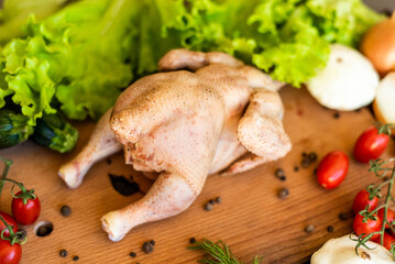 The chicken ready for cutting lies on a wooden board. Next to the chicken there are vegetables,...