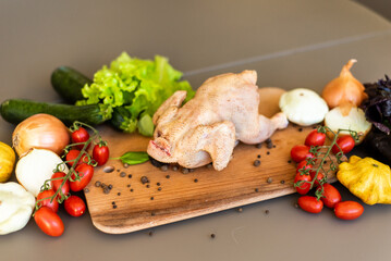 The chicken ready for cutting lies on a wooden board. Next to the chicken there are vegetables,...