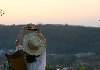 A long-haired female tourist in a straw hat sits on a brown chair using a mobile phone to photograph the beauty of the sunset. Evening atmosphere with mountains and sky. Holiday vacation concept.

