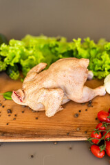The chicken ready for cutting lies on a wooden board. Next to the chicken there are vegetables,...