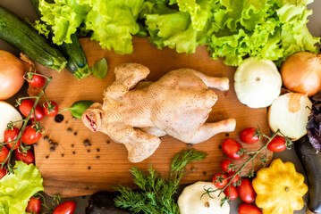 The chicken ready for cutting lies on a wooden board. Next to the chicken there are vegetables,...