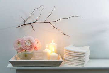 Three pale pink ranunculus in a transparent round vase and candles on the white windowsill. Rolled...