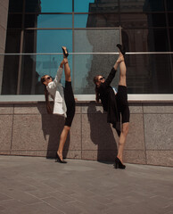 Fototapeta premium two sports young girls in trendy suits are standing fashion in stretching poses vertical tightening on the street modern building background at sunny day. fashion concept, free space