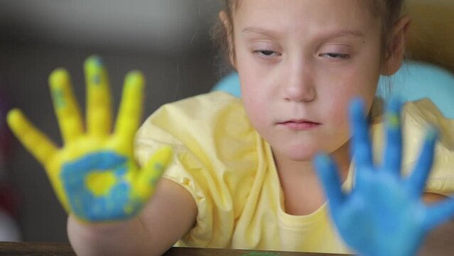 A sad child girl shows her hands painted in the colors of the Ukrainian flag, yellow and blue. Little Ukrainian patriot. Stop the war in Ukraine. Military conflict between Russia, Ukraine and the US