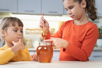 Happy children eating honey from a jar at home in the kitchen. funny girls lick honey from a spoon. Healthy organic food sweets for kids