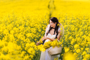 Fototapeta premium beautiful woman collects yellow rapeseed flowers in a large basket. A girl in a white dress and a big hat