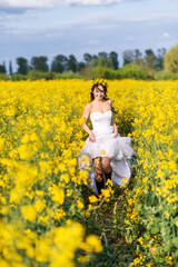 Fototapeta premium young beautiful woman runs through a field with yellow flowers. The bride in a long white dress and boots in a rapeseed field.