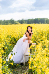 Fototapeta premium woman in a long white dress and black boots is sitting in a rapeseed field . The girl enjoys nature and freedom