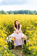 Fototapeta premium woman in a long white dress and black boots is sitting in a rapeseed field . The girl enjoys nature and freedom
