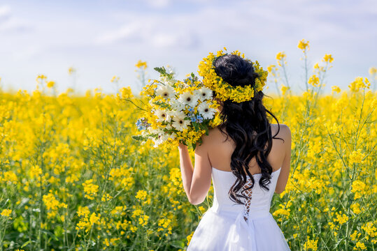 Portrait of a beautiful woman with a wreath of yellow flowers on her head. A girl with a bouquet in a rapeseed field. Photo from the back