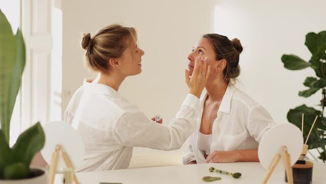 Cheerful young fair-skinned girl applies organic day cream to adult woman while sitting in bright room. Two ladies in shirts laugh and feel happy. Natural beauty, skin care concept