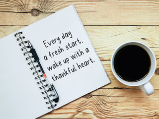 Open notebook with text "Every day is a fresh start, wake up with a thankful heart." and a cup  of coffee on wooden background.