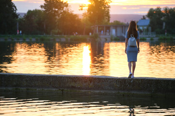 Fototapeta premium Back view of lonely young woman standing alone on lake shore enjoying warm evening. Wellbeing and relaxing in nature concept