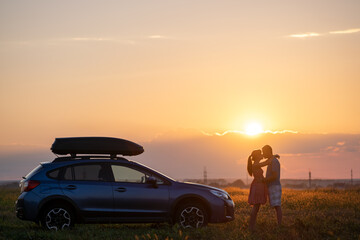 Fototapeta na wymiar Happy couple spending time together near their SUV car during honeymoon road trip at warm summer evening. Young man and woman enjoying road trip travelling by vehicle in nature
