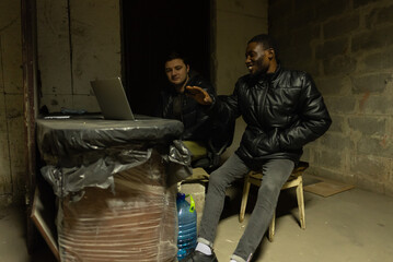 Fototapeta na wymiar Caucasian guy and black guy are hiding in a basement during a bombardment and having a talk near laptop