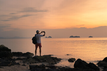 A man on the beach shoots a colorful sunset on a smartphone.Spectacular sunset sky over sea and islands.