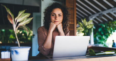 Portrait of millennial female IT professional with modern laptop device looking at camera during time for creating graphic design, skilled woman with digital netbook posing at terrace table