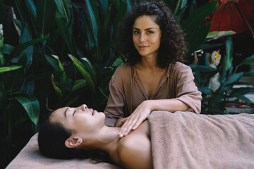 Fototapeta na wymiar Portrait of Caucasian woman doing professional Thai massage to Asian female client enjoying technique of procedure, cosmetologist doing rejuvenation for stretch skin and looking at camera during seans