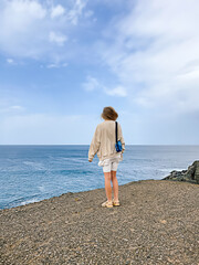 One young woman seen from back wearing white and beige clothes standing on a rock cliff on a windy day on blue sky and sea background. Travelling concept. Well Being and peacefulness.