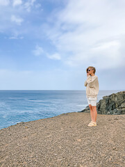One young woman seen from back wearing white and beige clothes standing on a rock cliff on a windy day on blue sky and sea background. Travelling concept. Well Being and peacefulness.