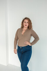 young plus size girl with red curly hair is standing in the beige bodysuit and blue jeans and smiling cute on the white wall background. fashion concept, free space