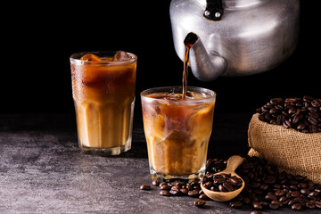 Pour coffee, iced latte into a glass. Add milk or cream along with roasted coffee beans. Set on a wooden table at a coffee shop on a dark black background.
