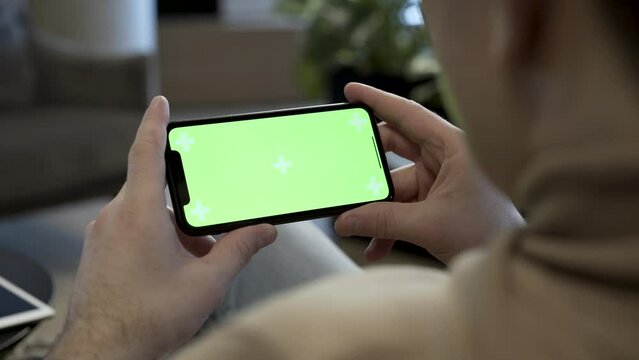 Young man holding mobile smartphone in landscape position with his fingers. He is looking at the screen watching pictures or video. Display is with green screen which is perfect for chroma keying.