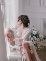 young girl with black short hair in white boudoir robe is sitting in profile near huge window on the white wall background with flowers. lifestyle concept, free space