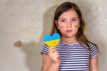 Stop war in Ukraine concept. Little Ukrainian blond girl, with the flag of ukraine in the heart in a ruined old house. Save the children, close the sky of Ukraine, humanitarian aid disaster concept