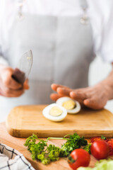 The cook in the kitchen slices a hard-boiled egg with a knife on a wooden board. Preparation of eggs and their processing. Home atmosphere in the kitchen. Next to lay greens and kitchen utensils.