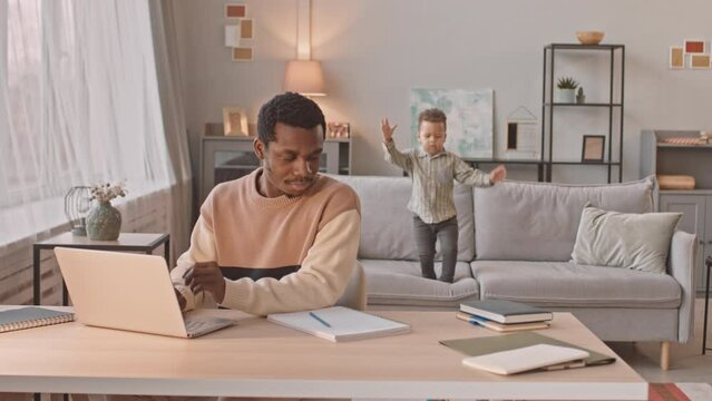 Medium slowmo of Black man using laptop while working from home, staying at home during lockdown with his toddler son playing in background