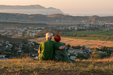 An elderly couple sits on a mountain with their backs with a beautiful view of the mountains and the sea in the distance.